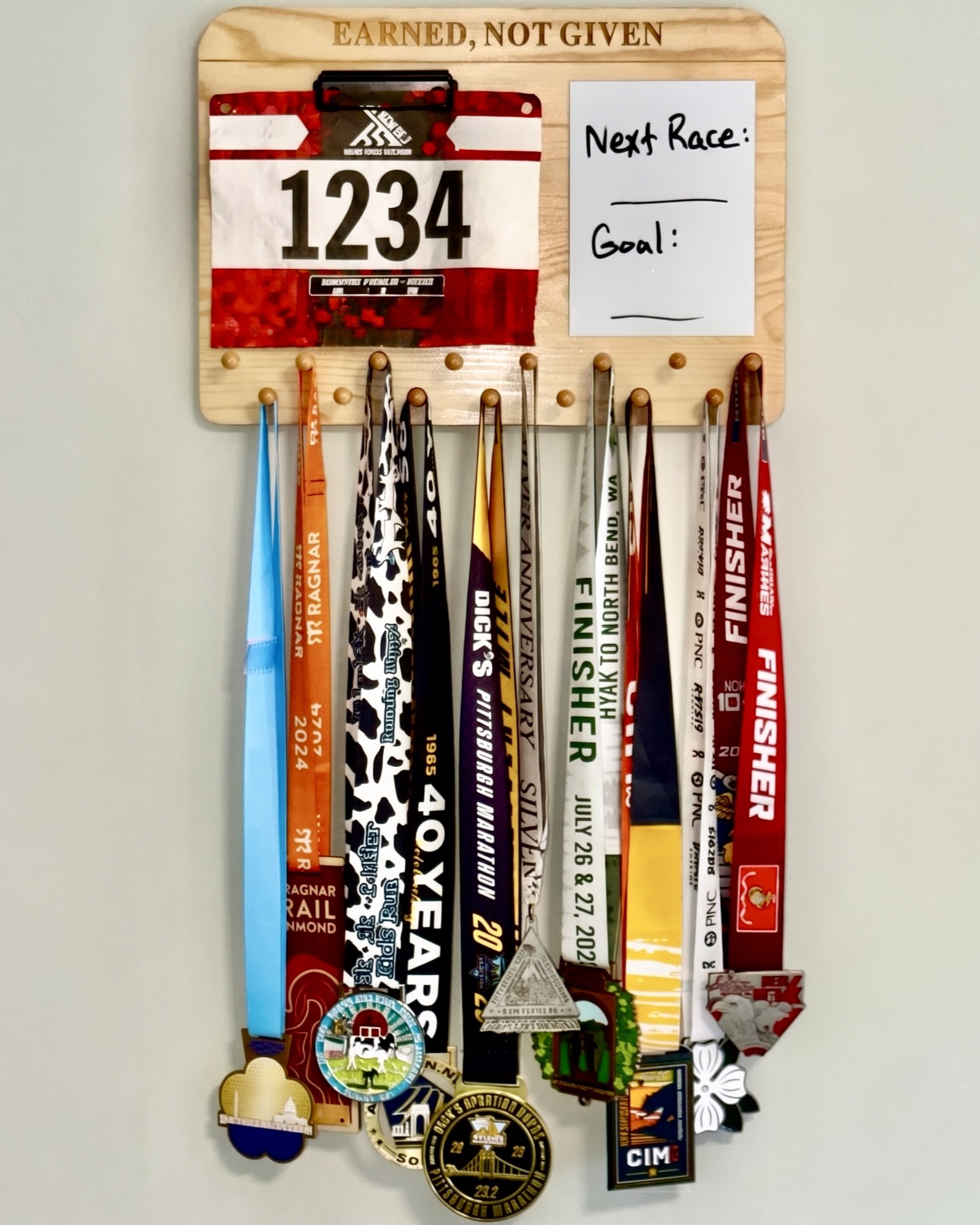 Solid wood medal hanger display holding race bibs and a full collection of race medals beside a goal whiteboard